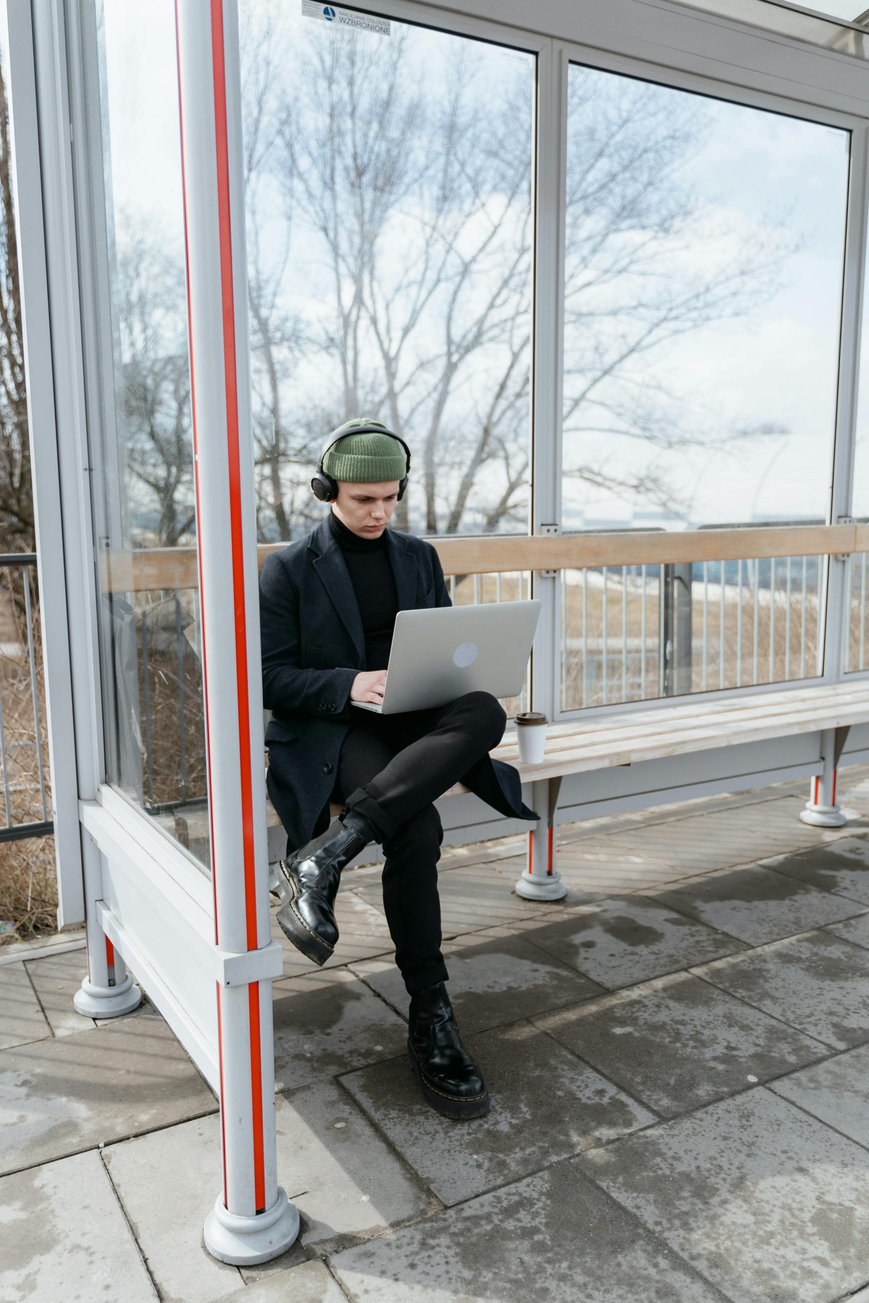 A person in a black coat sits at a bus stop, working on a laptop on a winter day.